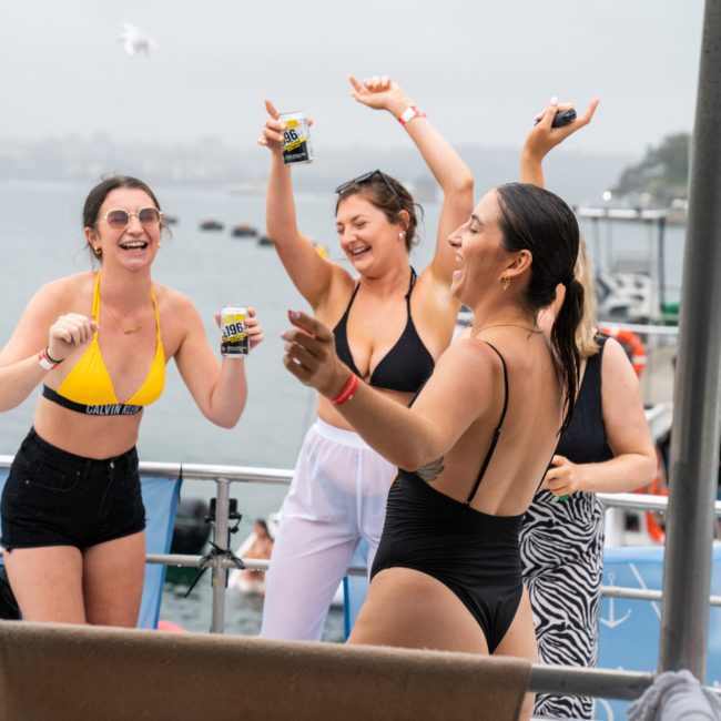 Four women in swimwear smile and dance on a boat with water and other boats visible in the background on a cloudy day, illustrating the joy of a Sydney boat party hire.