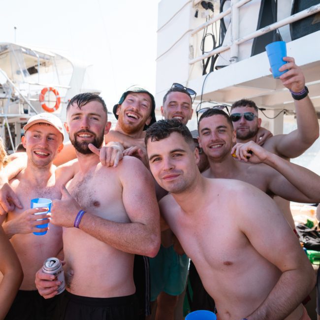 A group of people are gathered closely together on the deck of a boat, holding drinks and smiling towards the camera, enjoying what seems to be a corporate boat event in Sydney.