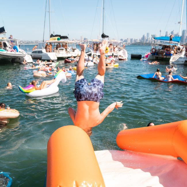 A person wearing swim trunks is caught mid-dive headfirst into the water from an orange inflatable float, surrounded by boats and other people on inflatable floats, with a stunning city skyline visible in the background. Enjoy moments like these with a luxury yacht hire in Sydney for an unforgettable experience.