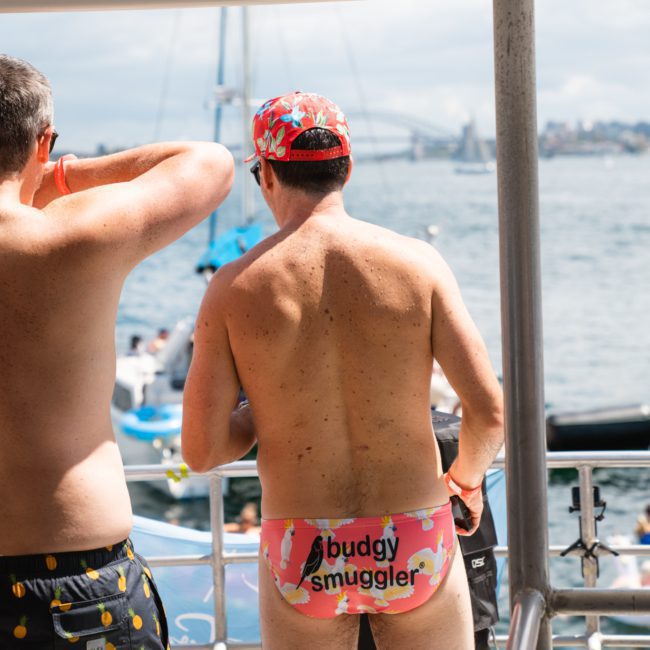 Two individuals in swimwear on a boat deck; one wears a cap and patterned underwear with "budgy smuggler" written on it, while the other is shirtless. A body of water and boats, including luxury yacht hire Sydney vessels, are visible in the background.