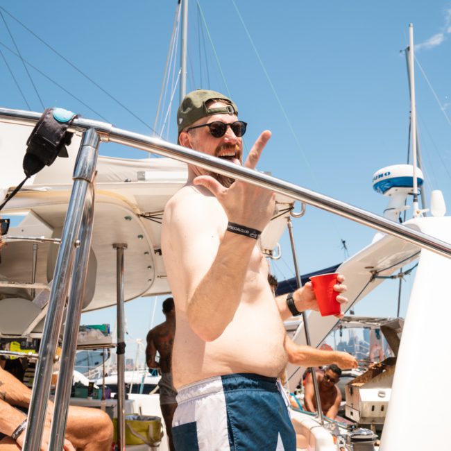 Description: A group of men socializing on a boat under a clear sky. One man, holding a red cup, is gesturing and wearing shorts and a cap. Another man is seated with a man bun. Various boat equipment is visible, making it ideal for those considering luxury yacht hire Sydney or private yacht charter Sydney Harbour.