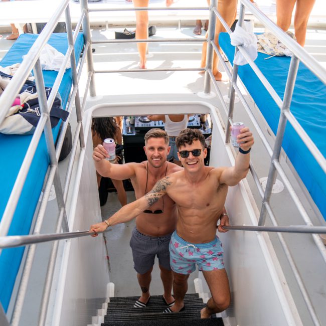 Two men in swim trunks and sunglasses are standing on the stairs of a boat, holding drinks, and smiling at the camera. Other people are in the background on the deck of this luxury yacht hire Sydney.