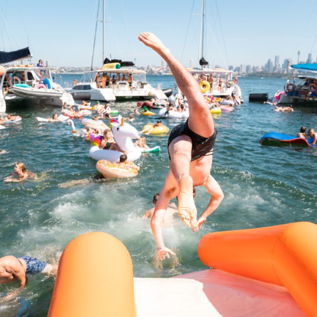 A person in swim trunks is mid-dive off an inflatable slide into a crowded water area. People float on various inflatables, and boats anchored in the background hint at the lively atmosphere of a Sydney boat party hire on a sunny day.