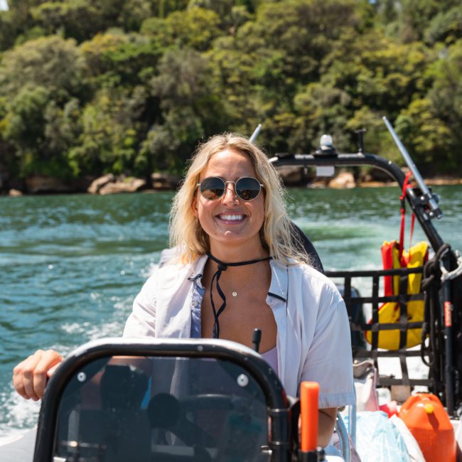 A woman wearing sunglasses and a white shirt smiles while steering a boat on the water with a wooded shoreline in the background, enjoying her DJ boat hire Sydney experience.