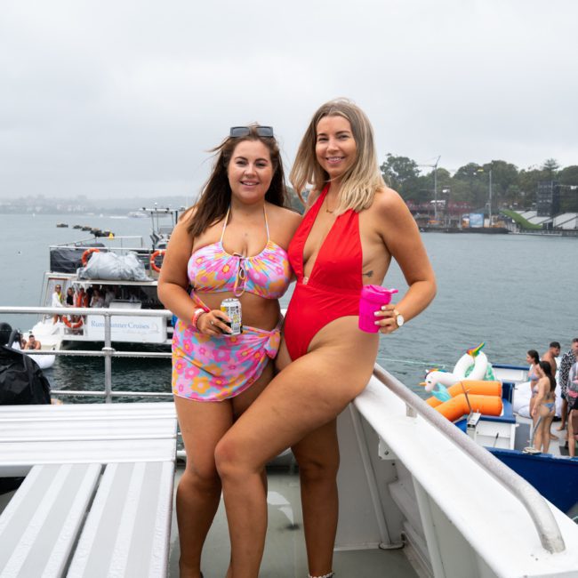 Two women in swimsuits pose on a luxury yacht with a scenic water backdrop. One wears a pink and purple suit, holding a can; the other wears a red suit, holding a pink cup.