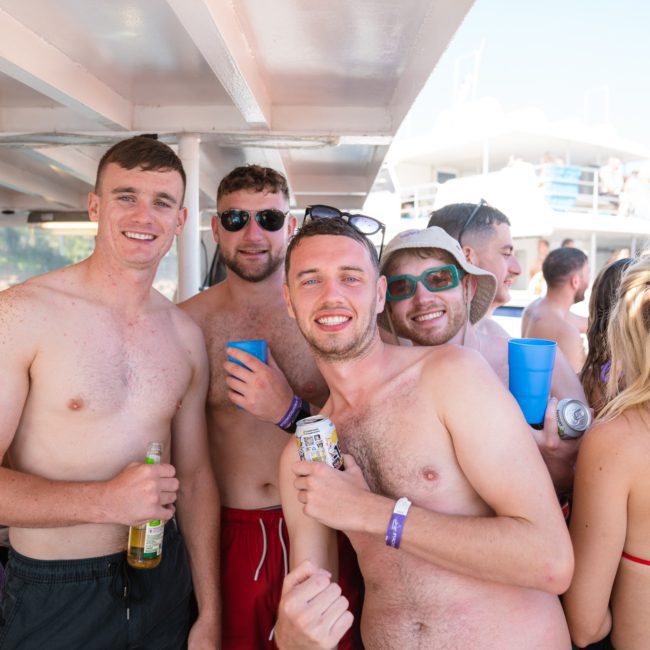 A group of young men and women are enjoying themselves on a boat. The men, shirtless and wearing swim trunks, are smiling and holding drinks. They are surrounded by other people in swim attire under a shaded area of the boat adorned with nautical decor. The mood is lively and cheerful.