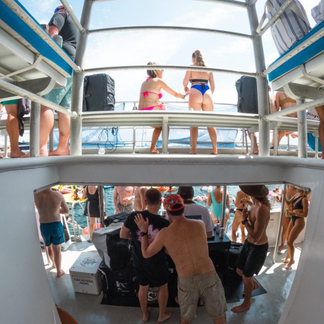 A group of people in swimwear socialize on a multi-level catamaran party Sydney under a clear sky. Some are near the railings, and others interact near a black table in the center. Water is visible in the background, creating an ideal setting for fun and relaxation.