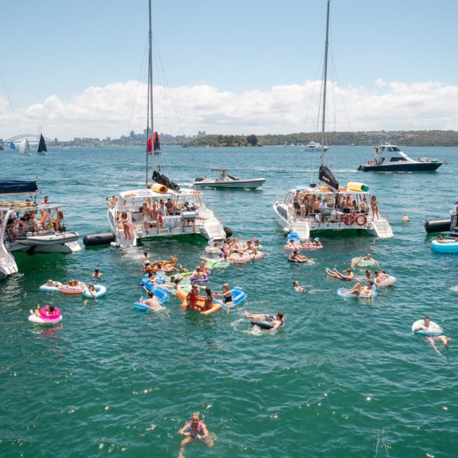A group of people on boats and floaties enjoy a sunny day on the water, with a city skyline in the background—a perfect scene for a Sydney boat party hire.