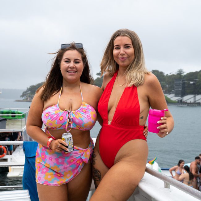 Two women in swimsuits stand on a boat holding drinks, with a group of people and a scenic water background visible, enjoying a catamaran party in Sydney.