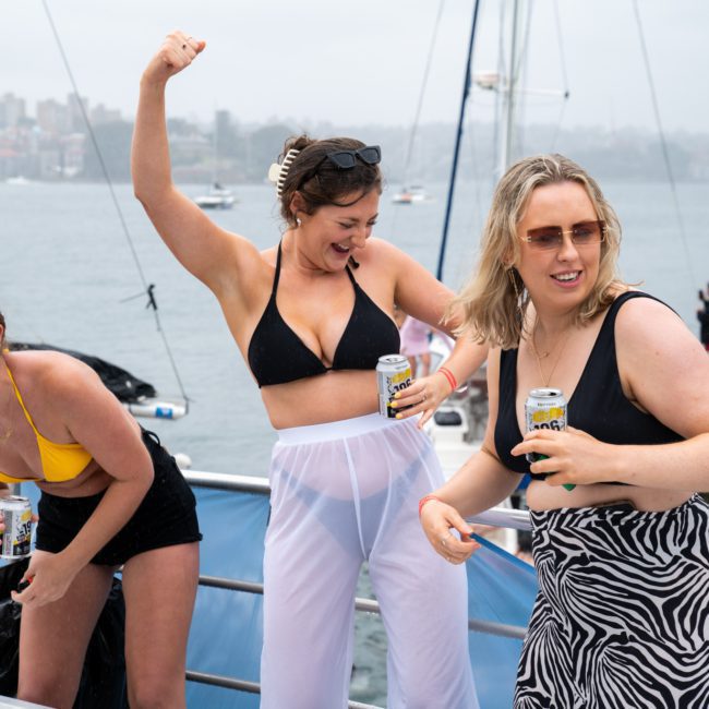 Three women in swimwear and boat attire are holding drinks and dancing on a luxury yacht. The background shows a cloudy sky and waterfront with other boats visible, adding to the vibrant atmosphere of what could be an unforgettable Sydney boat party hire.