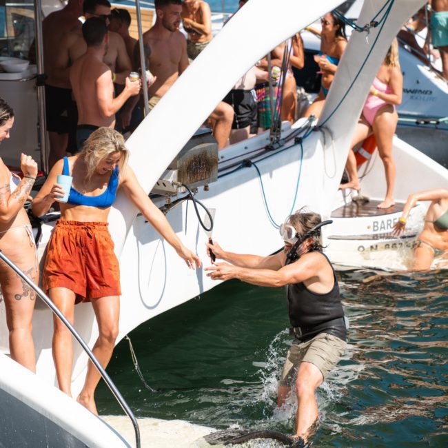 People socialize on a luxury yacht hire in Sydney. A person in the water wearing a snorkel mask reaches out to another who is standing on a narrow platform extending from the boat. City buildings are visible in the background, making it perfect for corporate boat events or a Sydney boat party hire.