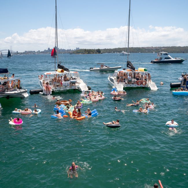 A group of anchored boats with people swimming and floating on inflatable tubes in clear, blue water under a sunny sky, perfect for enjoying a DJ boat hire in Sydney or perhaps one of those exclusive private yacht charters on Sydney Harbour.