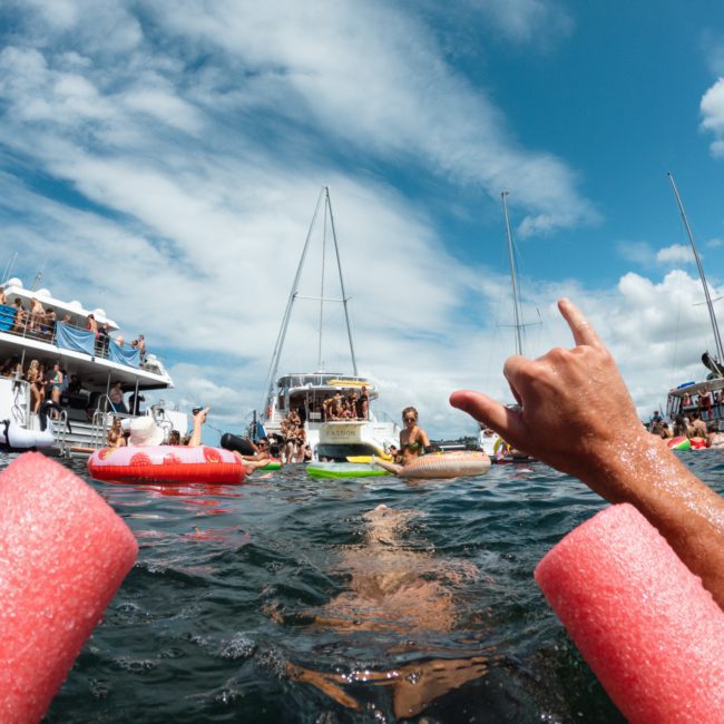 People on boats and in the water enjoying a sunny day, with some using pool noodles and inflatable floats. In the foreground, a hand is making a "hang loose" gesture—typical scenes from a catamaran party Sydney loves to host.