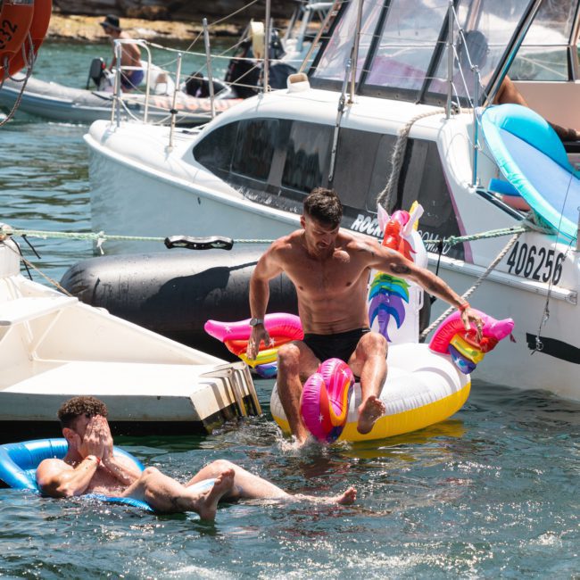 Two men enjoy a sunny day on the water, one sitting on an inflatable unicorn while the other laughs in the water. Nearby, boats are docked, perfect for a private yacht charter on Sydney Harbour.