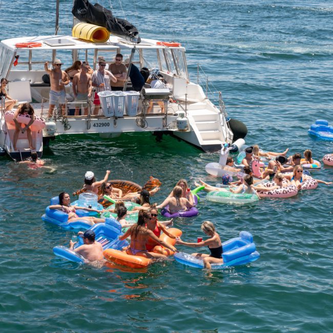 A group of people on inflatable pool floats enjoy the water near a boat on a sunny day. The boat is anchored, and more people are gathered on the deck. It looks like a perfect setting for a Luxury yacht hire Sydney event.