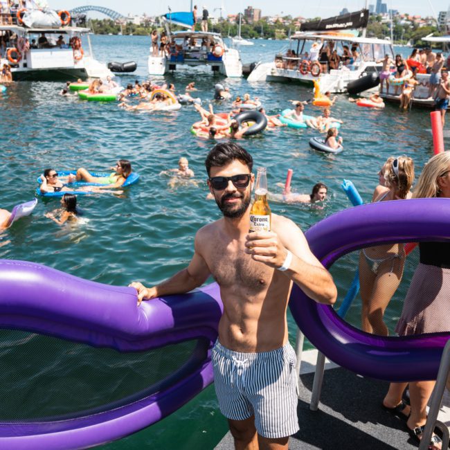 A man wearing sunglasses and striped swim trunks holds a bottle of Corona beer on a boat. Behind him, numerous people enjoy the water on inflatables with boats in the background, epitomizing the vibrant atmosphere of a Sydney boat party hire.