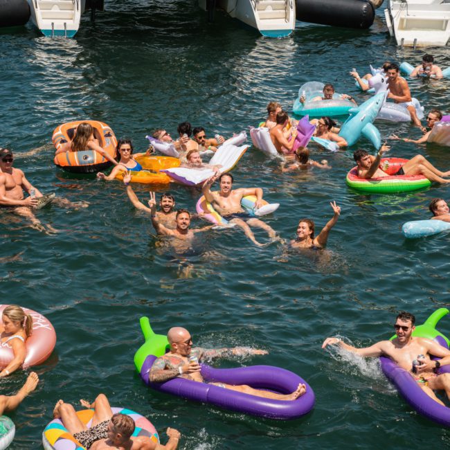 A lively group of people enjoys a sunny day in the water, floating on various colorful inflatables, including unicorns, donuts, and flamingos. Some wave at the camera while others relax and chat. Paddleboards and boats are docked in the background.