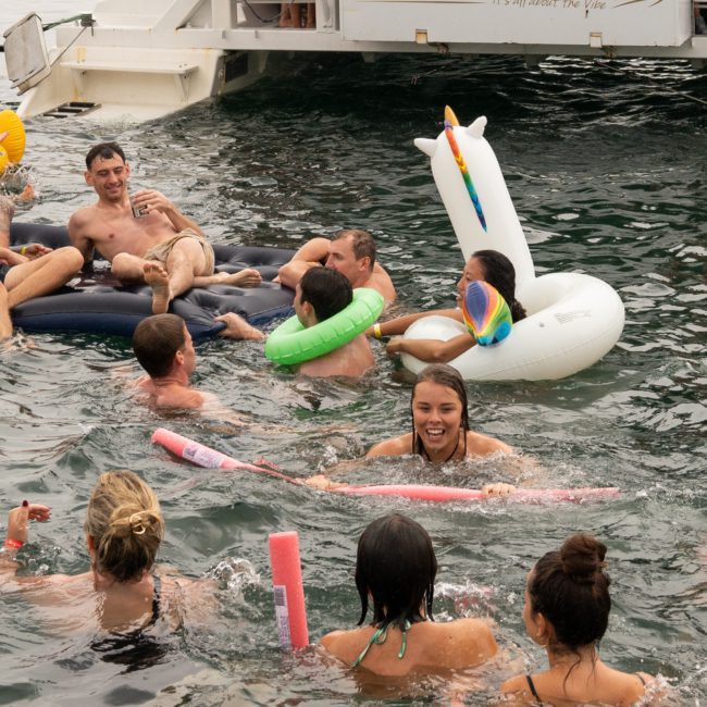 A group of people are swimming and relaxing on inflatables in the water near a luxury yacht. Some are chatting, and one person is on a unicorn float. It appears to be a social gathering enhanced by DJ boat hire Sydney services.