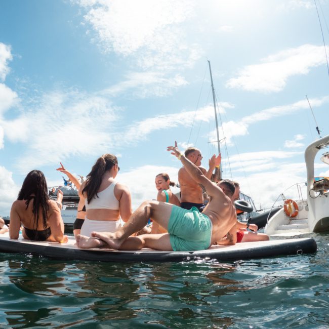 A group of people on an inflatable float in the water, surrounded by boats, under a sunny sky, enjoys the vibes of a catamaran party in Sydney.