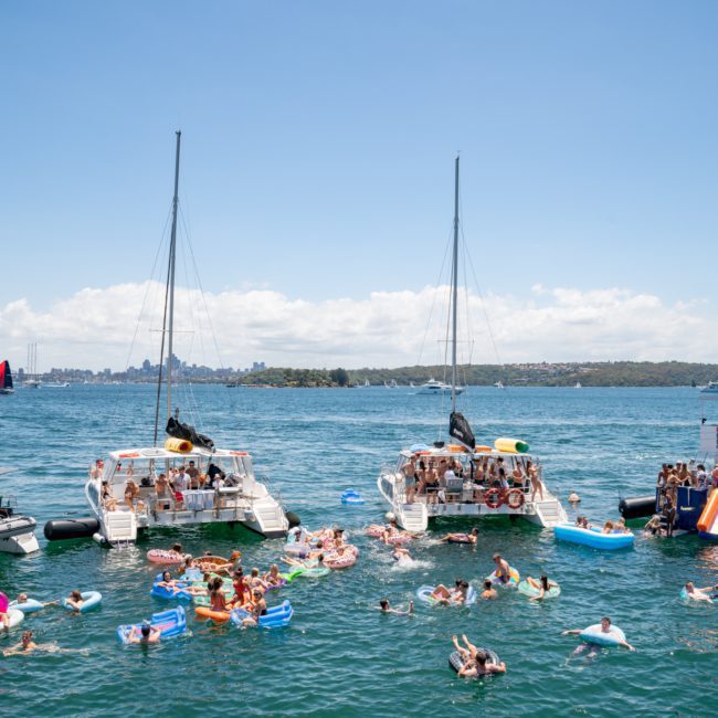 Private yachts and boats gathered on the water with numerous people swimming and floating on inflatables in a sunny, scenic setting.