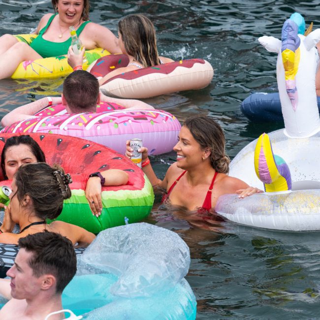 People are floating on colorful inflatable pool toys, including a watermelon, a donut, a tire, and a swan, in a body of water. Some are holding drinks and chatting with each other. Nearby, there's an exciting Sydney boat party hire featuring lively DJ music to elevate the experience.