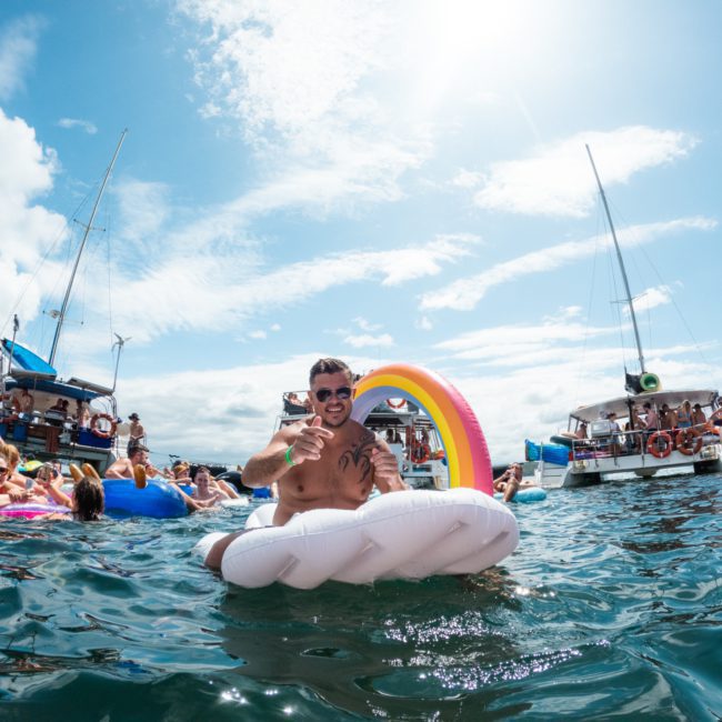 People enjoying a sunny day on the water with colorful inflatables and boats in the background. Two men are smiling on a white cloud-shaped float with a rainbow arch, adding to the vibrant atmosphere of a luxury yacht hire in Sydney.