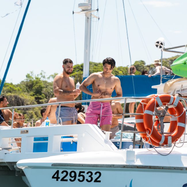 Two men standing on the deck of a boat with the number 429532, surrounded by other people. Enjoying a Sydney boat party hire, the boat is on the water near a wooded shoreline.
