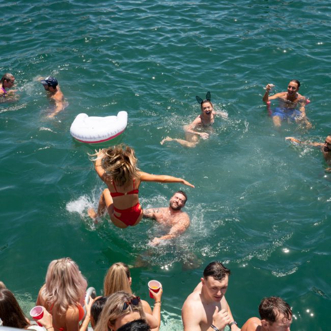 Group of people swimming and enjoying water activities in a sunny outdoor setting. A person in a red swimsuit is mid-air while jumping into the water from a luxury yacht hire in Sydney.