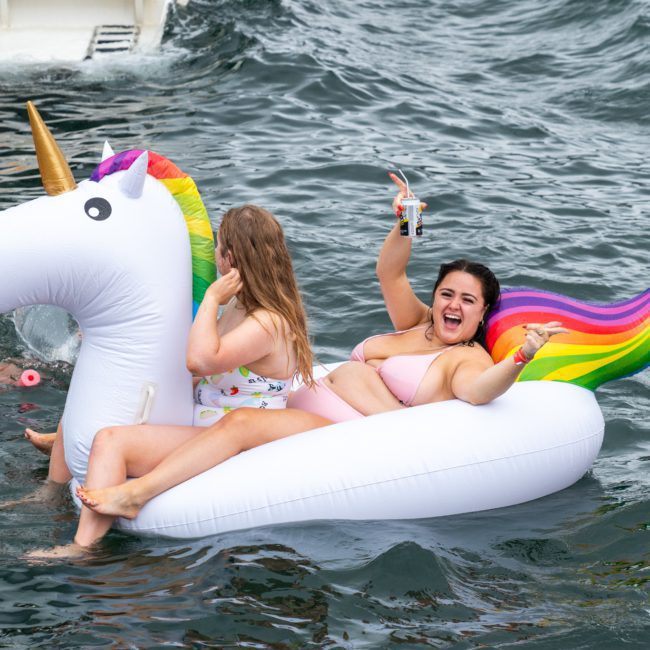 Two women on an inflatable unicorn float in the water, one holding a drink and waving, others swimming nearby. Consider opting for a Sydney boat party hire for more fun experiences like these, perfect for making lasting memories.