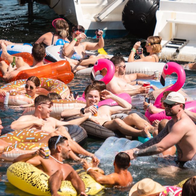 A large group of people relax and socialize on inflatable rafts and floats in the water, surrounded by flamingo and donut-shaped pool toys, near a docked private yacht charter Sydney Harbour.