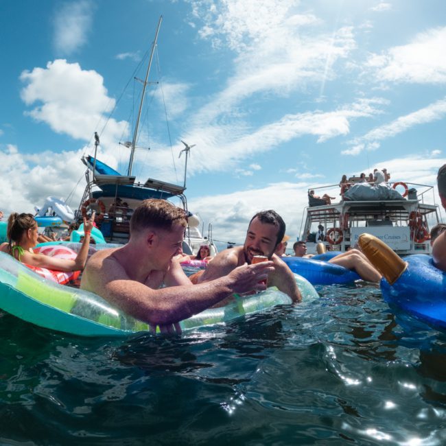 People relaxing on inflatables in the water, near boats, under a sunny sky. Some are engaged in conversation, while others sunbathe on the boats in the background. It's a scene straight out of a catamaran party Sydney experience.