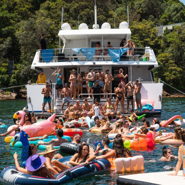 A large group of people are enjoying a sunny day at a lake. Many are on colorful inflatable rafts by a docked yacht, while others are swimming or standing on the yacht’s lower deck, embracing the essence of a Sydney boat party hire.