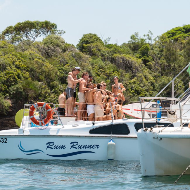 A group of people in swimwear gathers on a boat named "Rum Runner," docked near a forested shoreline. Another boat is partially visible with a few people on board and a life preserver on the side, hinting at the ideal setting for corporate boat events or Sydney boat party hire.