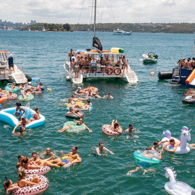 People enjoying a sunny day on boats and inflatables in the water, with a city skyline and islands in the background, perfect for a private yacht charter on Sydney Harbour.