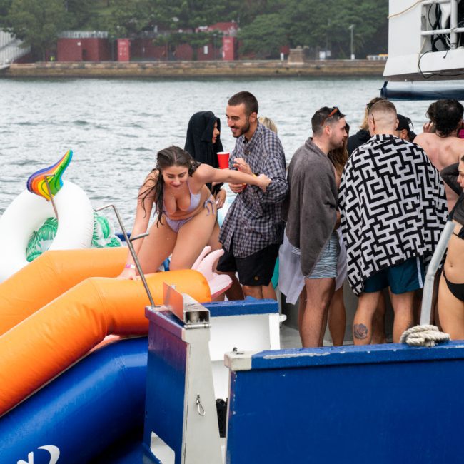 People enjoy a Sydney boat party, with some using an inflatable slide and others socializing in swimwear. Two unicorn floaties are visible in the water. The background shows a coastline with trees and buildings, perfect for a luxurious yacht hire experience in Sydney.