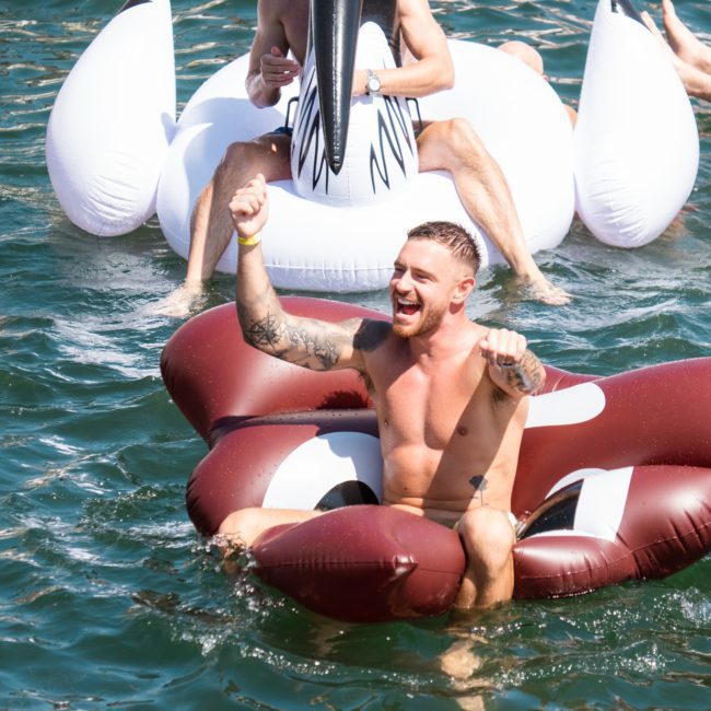 A shirtless man with tattoos floats in the water on an inflatable raft, smiling and raising his fists. Others on similar inflatables are visible in the background, enjoying what looks like a lively Catamaran party in Sydney.