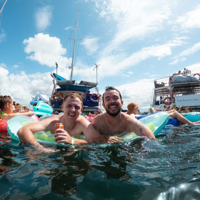 Two men smile while floating in the water on inflatable tubes at a vibrant Sydney boat party hire under a sunny sky.