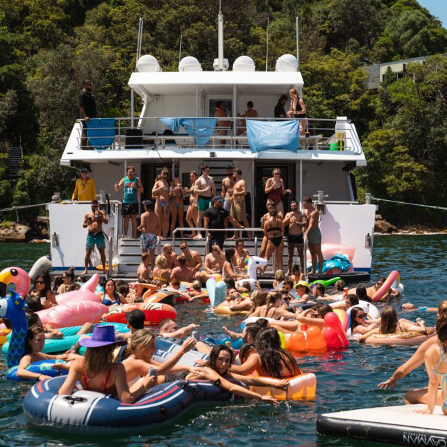 A large group of people on a boat and in the water with various inflatables, enjoying a lively Sydney boat party hire on a sunny day. Trees and a house are visible in the background.