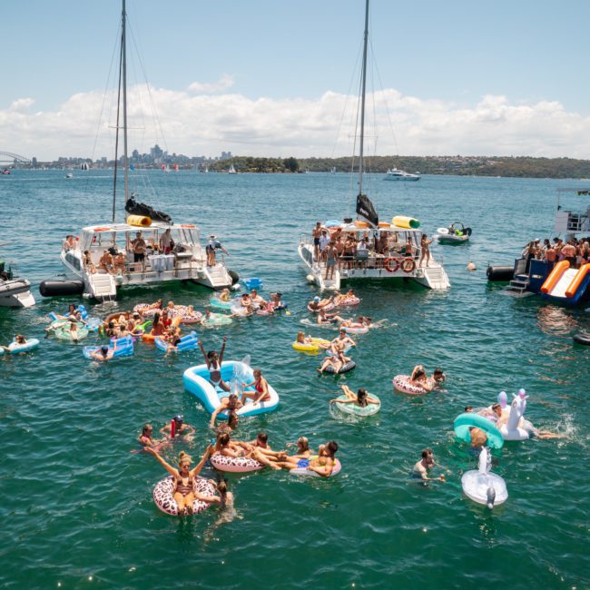 People are enjoying a sunny day on a lake, floating on inflatable rafts and swimming near boats docked together. It feels like a luxury yacht hire in Sydney with the perfect vibe for relaxation and fun.