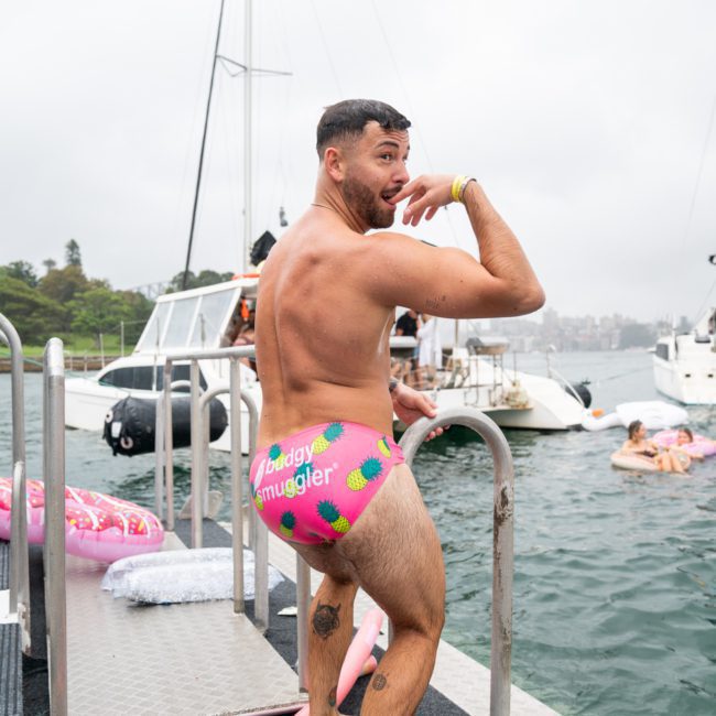 A man in patterned swimwear stands on a dock near the water, with boats and people in the background. He is looking over his shoulder and making a playful pose, possibly considering joining a DJ boat hire Sydney for an unforgettable experience.