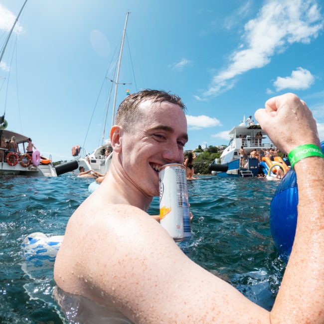 A man in a pool holding a can in his mouth and a blue float. Other people and several boats are visible in the background under a clear, blue sky, ideal for a Sydney boat party hire.