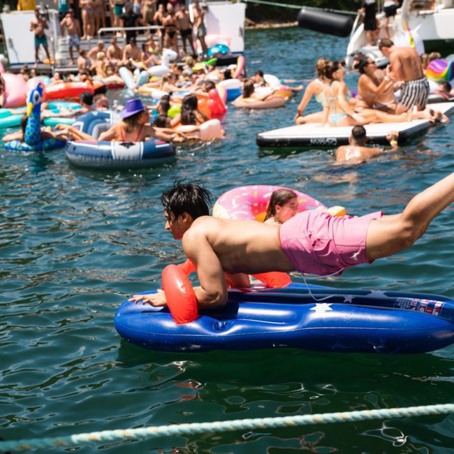 Numerous people on inflatables and in the water near boats at a party, with one man in pink shorts diving onto a float in the foreground, perfect for a Sydney boat party hire.