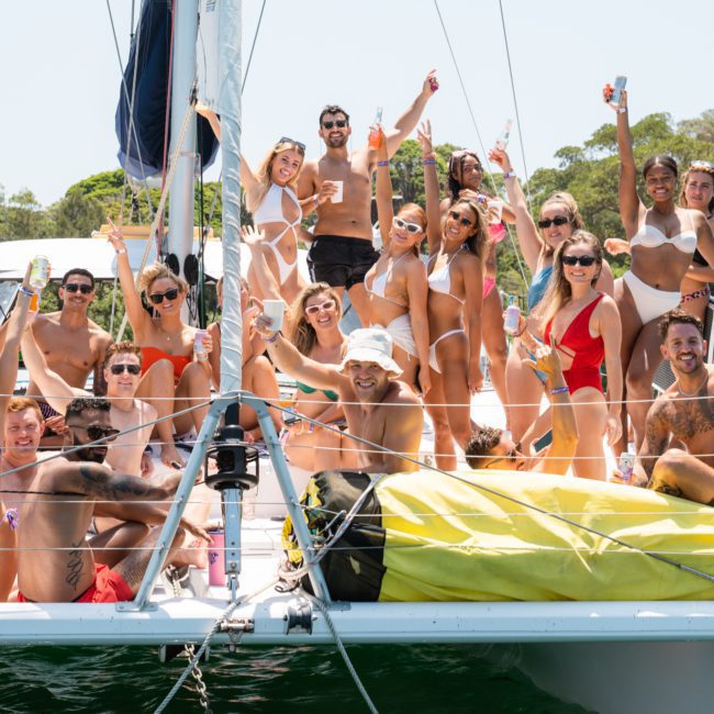 A group of people wearing swimsuits are gathered on a sailboat in a sunny location, smiling and raising their hands. A perfect scene for Sydney boat party hire, the background features more boats and trees.