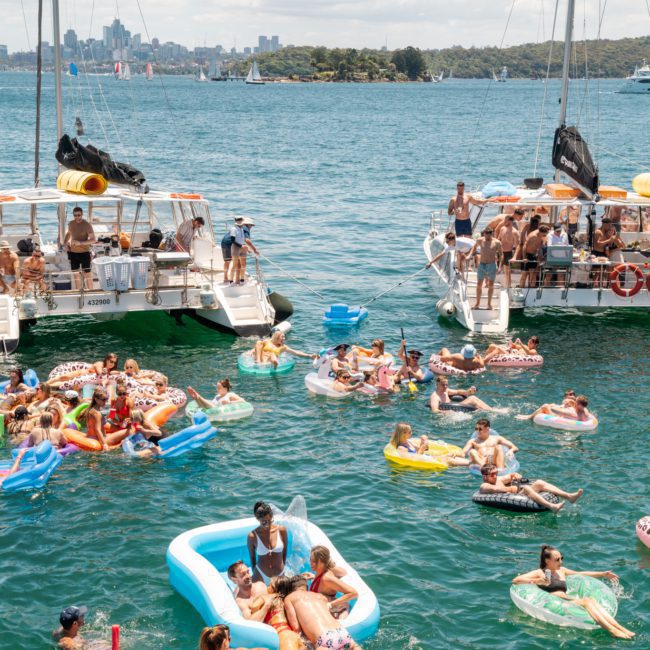 People enjoy a sunny day on the water, floating on inflatable pool toys near anchored boats. Some are swimming, while others are lounging and socializing. The city skyline adds to the charm of a Sydney boat party hire.