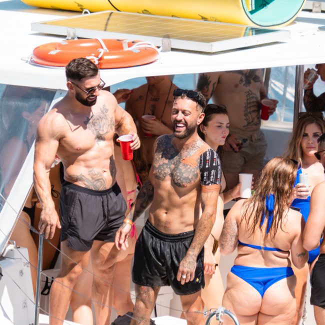 A group of people in swimsuits are socializing on a boat deck, enjoying a luxury yacht hire in Sydney. Several hold red cups, and some are laughing. In the background, a life ring and a yellow item are visible on the roof.
