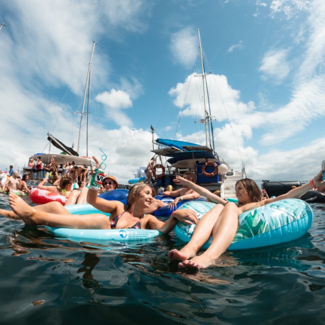 People are relaxing on inflatables in the water near a boat under a partly cloudy sky. Several boats, including luxurious yachts available for hire in Sydney, are visible in the background.
