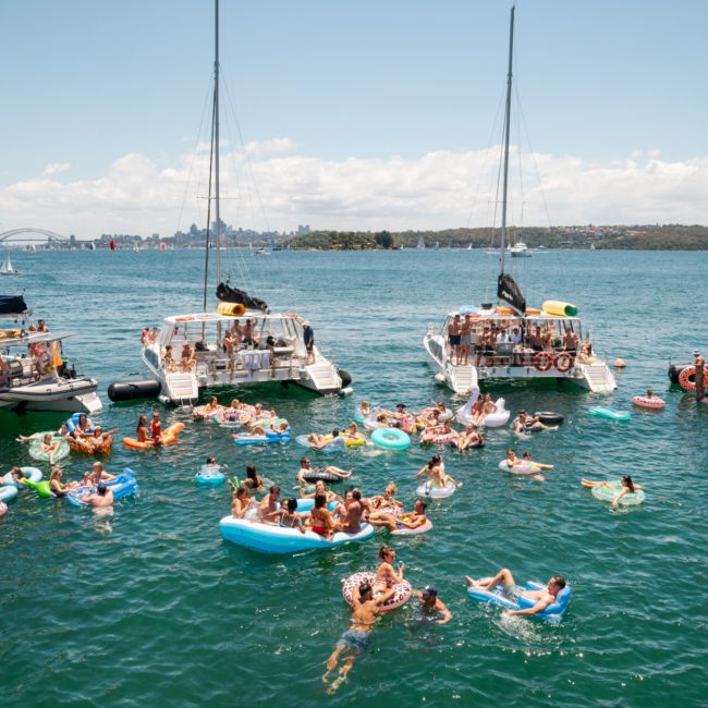 People on inflatable floats and boats enjoying a sunny day on a body of water, with a distant cityscape and bridge visible in the background. A private yacht charter Sydney Harbour adds an extra touch of luxury to the lively scene.