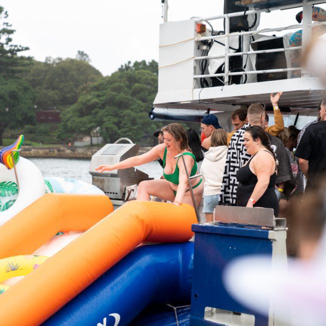 People are gathered on a boat's deck with an inflatable unicorn nearby, enjoying the vibrant atmosphere of a Sydney boat party hire. A person in a green swimsuit descends an orange slide into the water. Trees and shoreline are visible in the background, making it a perfect setting for corporate boat events Sydney.