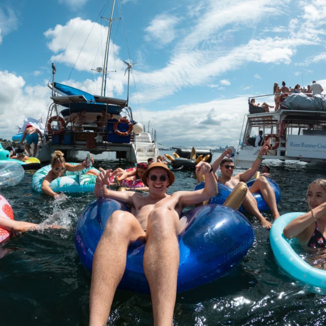 People floating on inflatable tubes in the water near boats on a sunny day. Some are smiling and posing for the camera. The sky is clear with some clouds, adding to the perfect ambiance of a luxurious yacht hire Sydney experience.