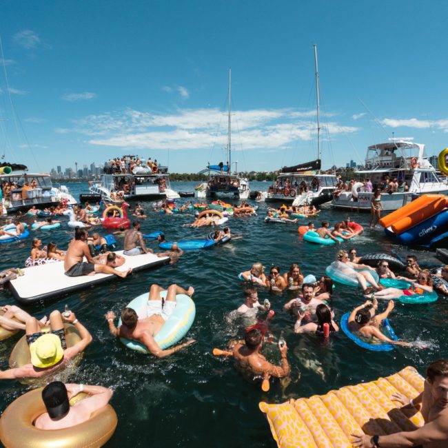 A crowded scene of people enjoying a sunny day on the water, floating on inflatables and swimming, surrounded by multiple boats. A DJ boat hire Sydney adds to the vibrant atmosphere with lively music.